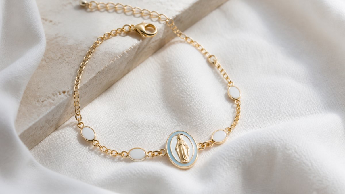 Close-up of a delicate gold-plated Miraculous Medal bracelet resting on soft white fabric with natural light highlighting the enamel details and medal centerpiece, showing elegant craftsmanship and sizing