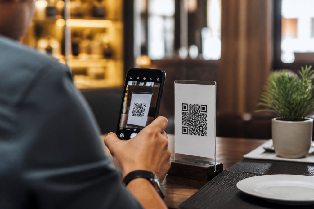 Cliente en un restaurante escaneando un código QR con su smartphone para acceder a un menú digital, con platos coloridos y atractivos visibles en la pantalla iluminada, ambiente de restaurante acogedor