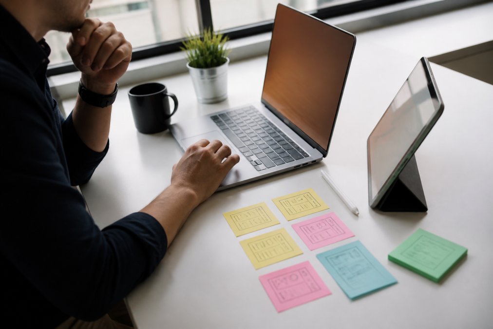 Persona trabajando en un portátil y tableta con interfaz de plataformas no-code y bocetos de aplicaciones en la mesa