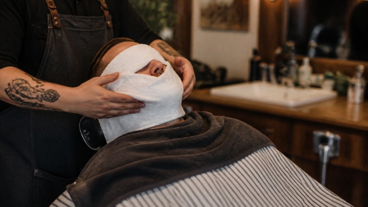 Man relaxing in a barber chair during his grooming rituals with a warm towel over his face