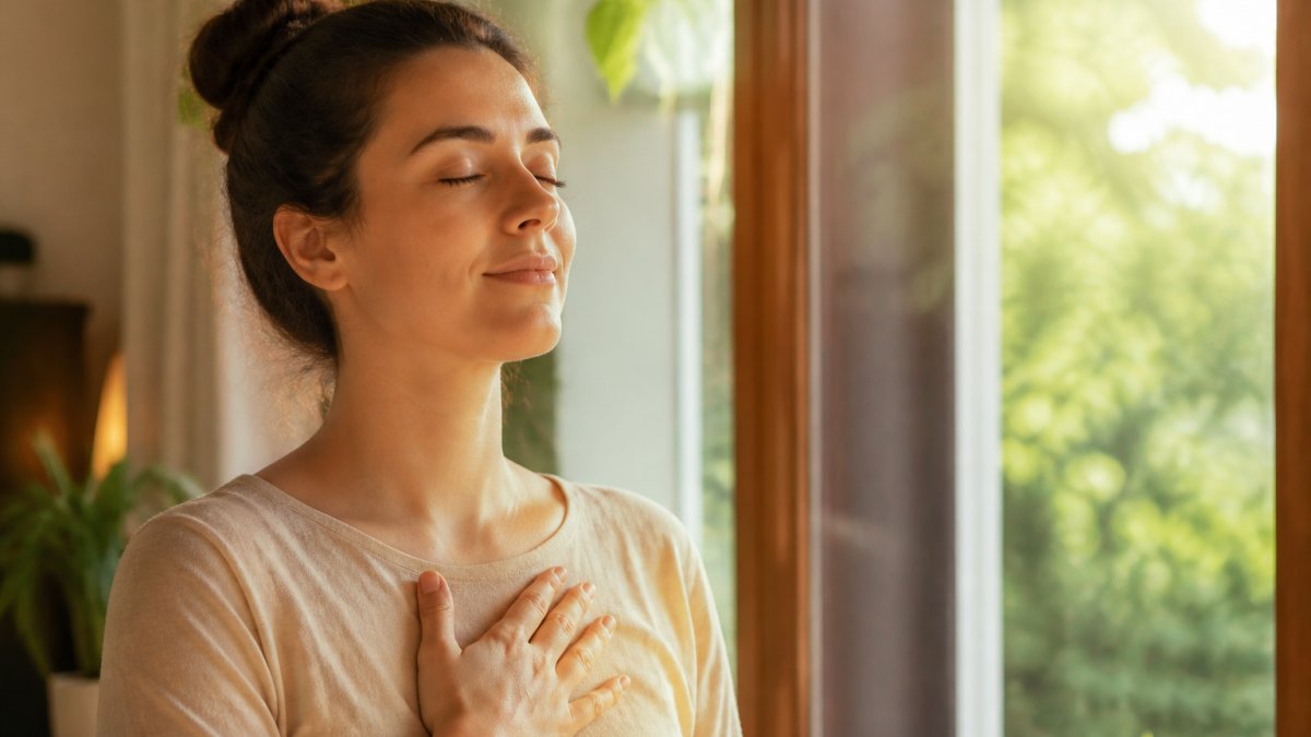 Woman sitting peacefully in natural sunlight near a window, demonstrating calm breathing with hand on chest, shoulders relaxed and eyes closed