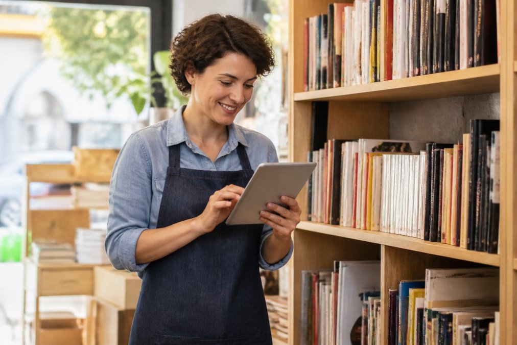 Libraia indipendente che sorride mentre controlla i livelli di scorte su un tablet davanti a uno scaffale di libri, luce naturale da una vetrina, atmosfera accogliente