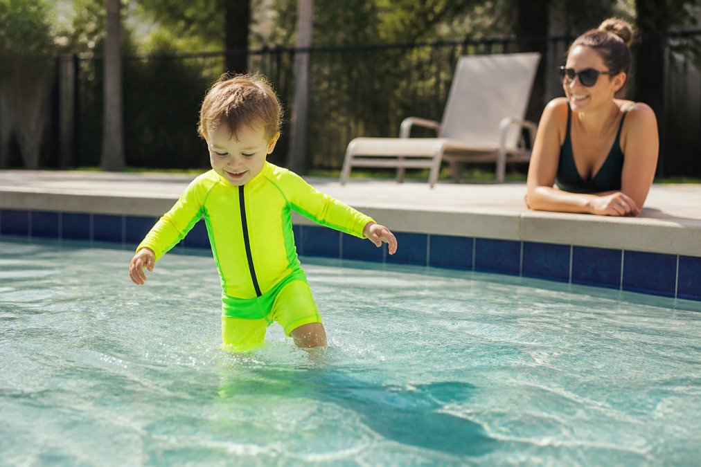 Toddler in a bright high-visibility swim set playing in a shallow pool while a parent watches to prevent causes of drowning.