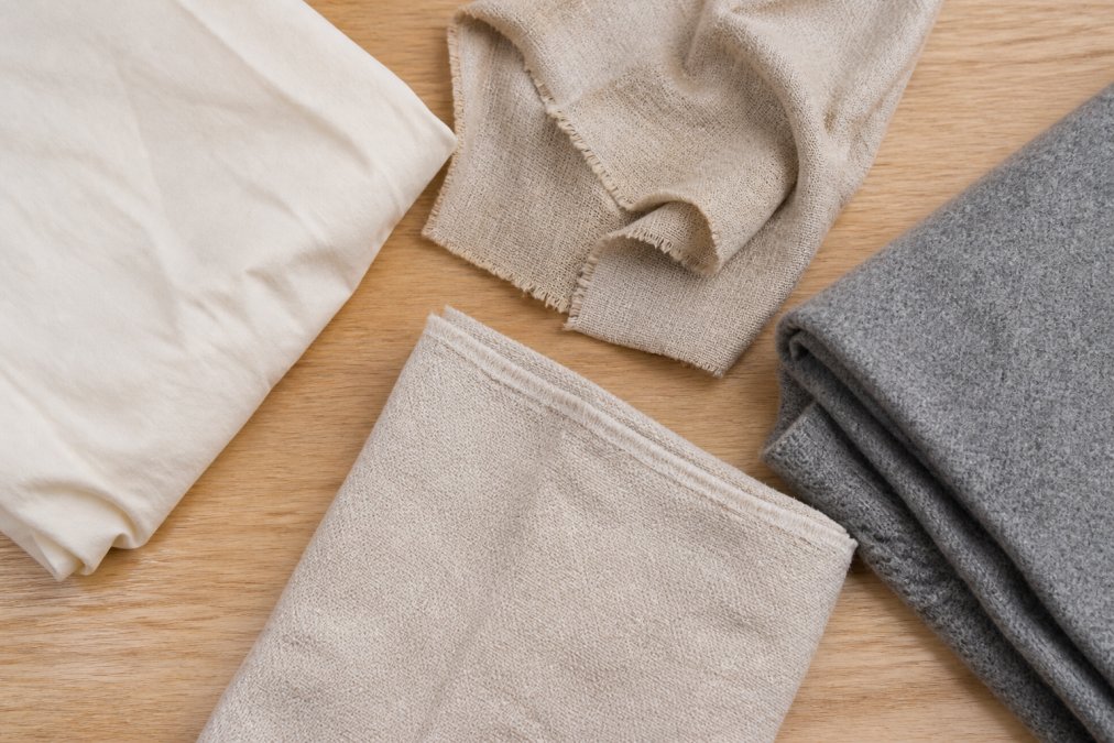 Overhead flat lay of natural fabric swatches including cream cotton, beige linen, undyed hemp, and folded gray wool arranged on a pale oak table in soft daylight
