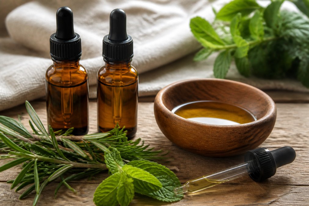 Close-up of essential oil bottles including rosemary and peppermint with glass dropper applicators next to a wooden bowl of golden carrier oil on a natural wood surface
