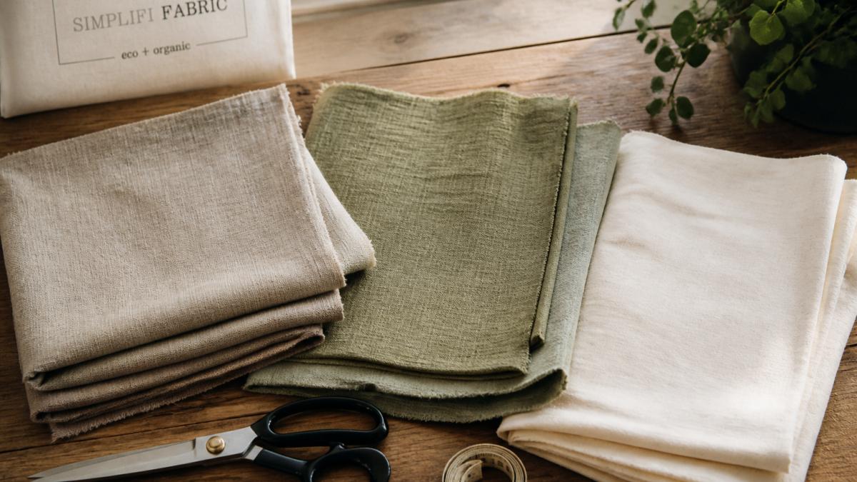Folded swatches of linen, hemp, and cotton on a wooden table with scissors and a measuring tape in soft light.
