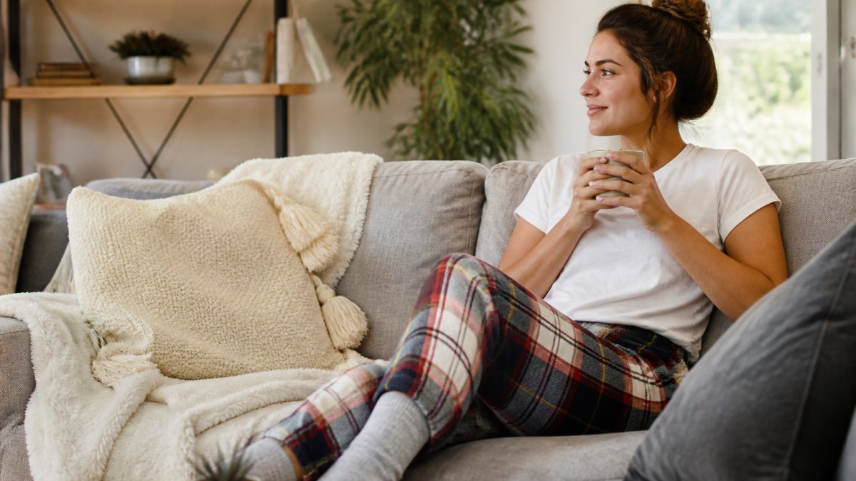 A woman relaxing on a couch in plaid joggers and a plain white T-shirt, holding a mug, with a cozy living room and natural light in the background