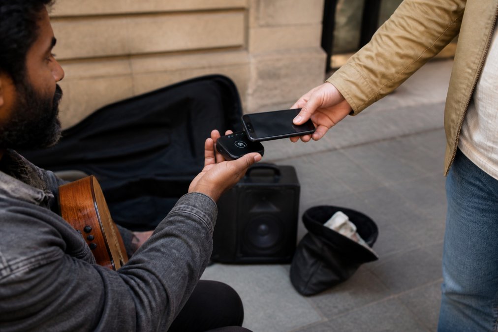 A supporter taps a smartphone on a street performer's NFC support display beside a guitar case and portable amplifier.