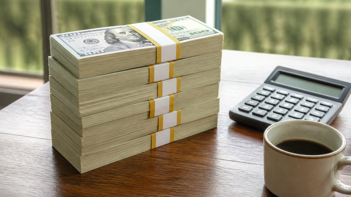 A stack of banded $100 bills on a wooden table next to a calculator and a coffee cup, shot in natural window light