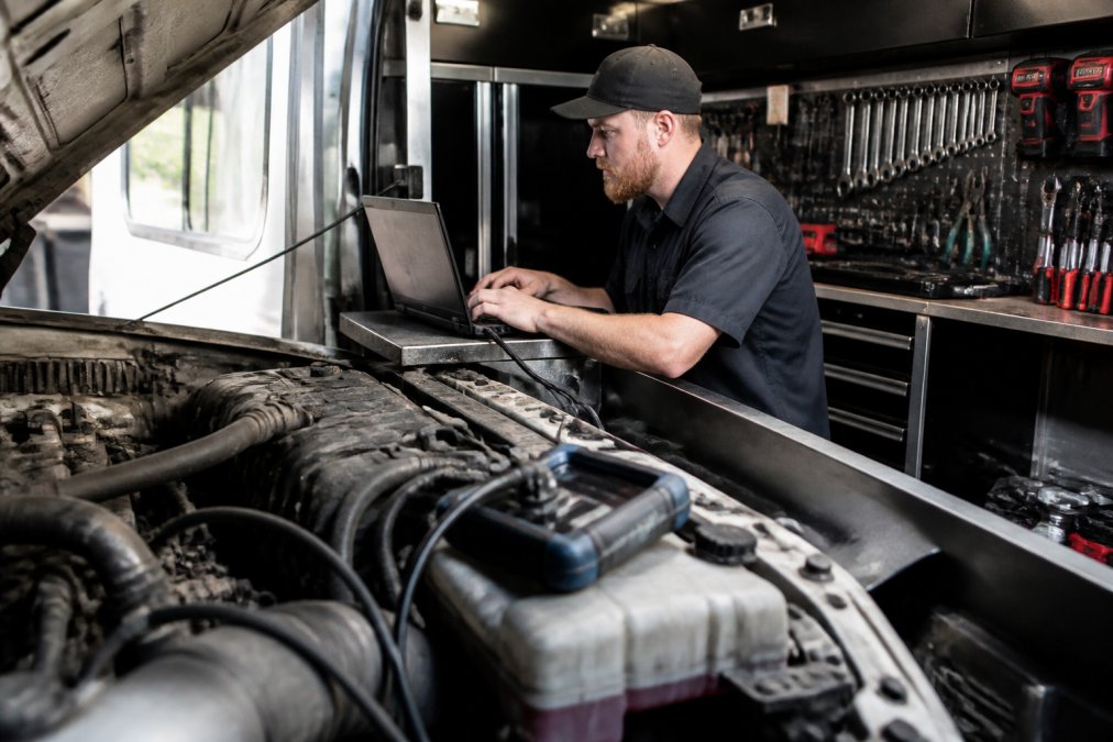 Professional diesel technician using laptop computer connected to diagnostic scanner inside mobile service truck, with engine bay visible and specialized tools mounted on truck walls