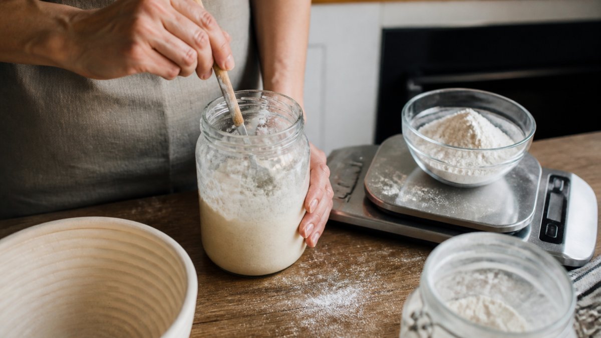 Baker stirring flour and water into a glass jar on a wooden counter
