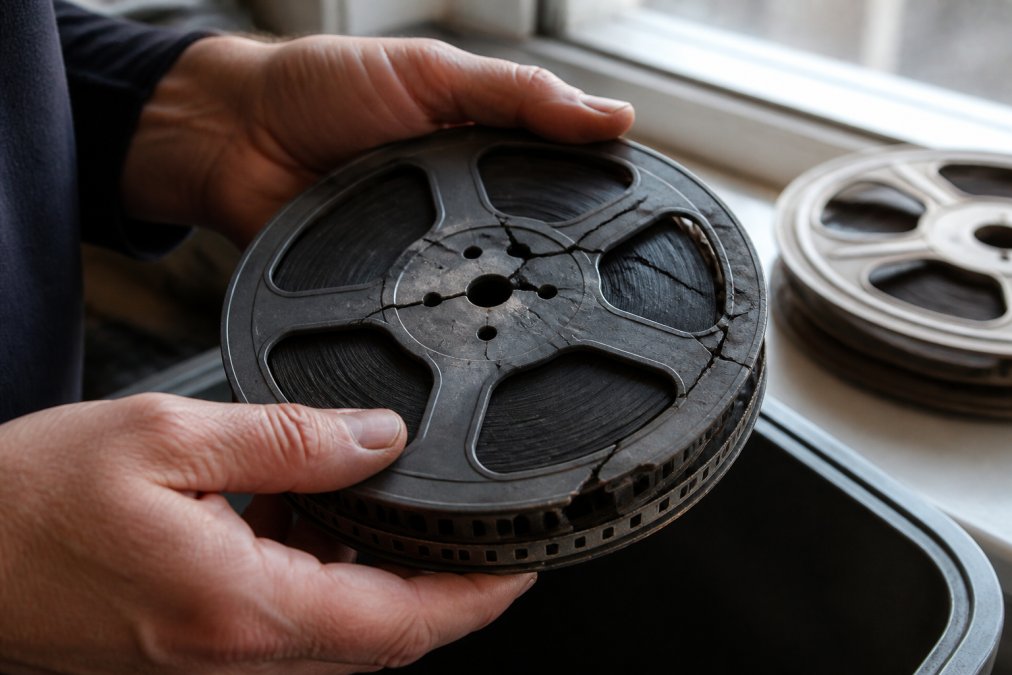 Close-up of a person's hands carefully holding a brittle, cracked 8mm film reel with visible sprocket hole damage, soft natural light from a window