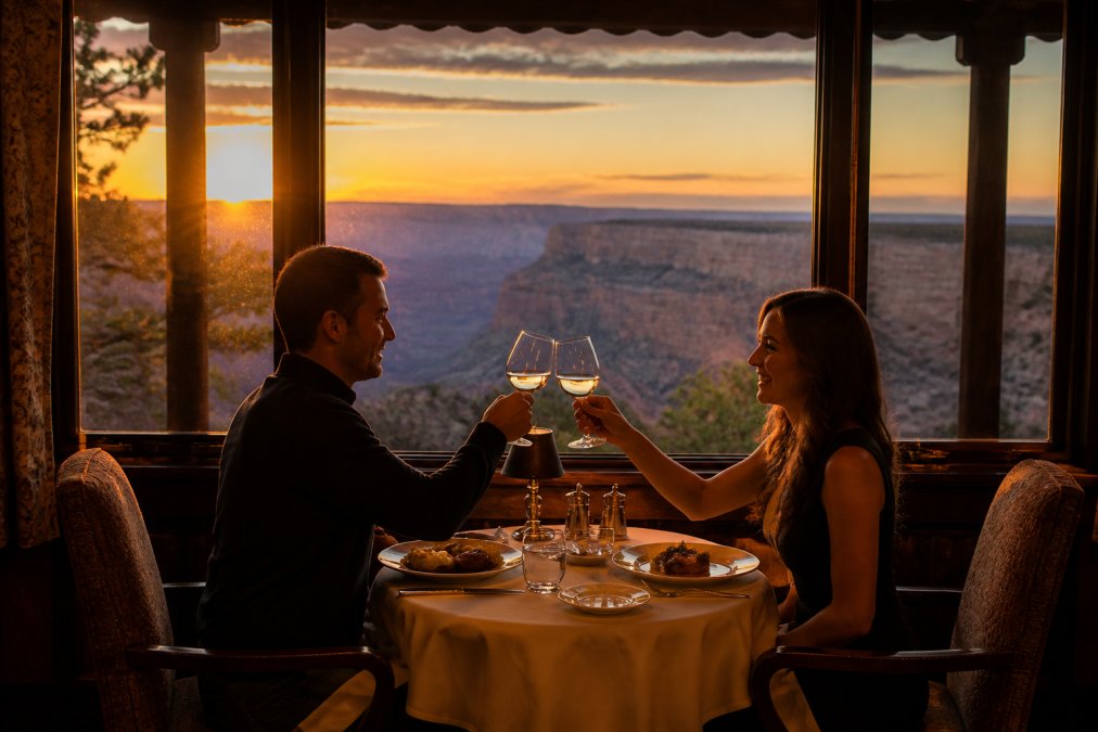 A couple toasting at a window table in a historic lodge dining room, golden evening light streaming in with a view of the canyon rim outside