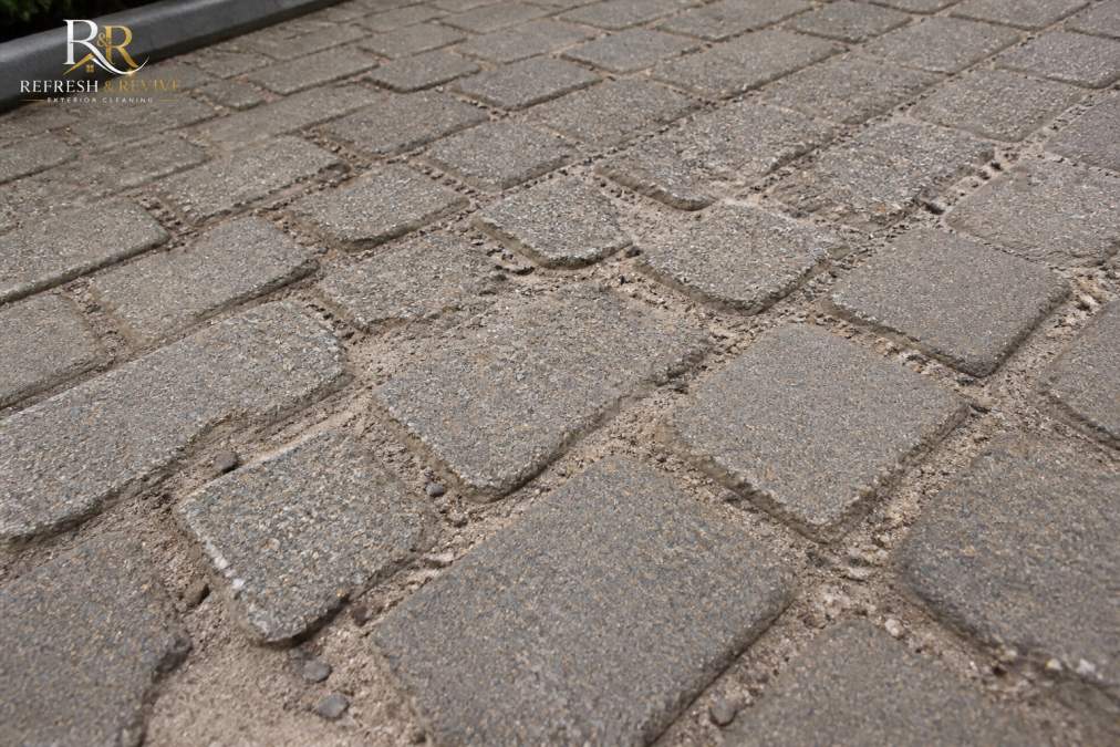 Close-up of a block paving driveway with eroded sand joints and a pitted, rough surface texture under overcast daylight, showing clear evidence of high-pressure washing damage