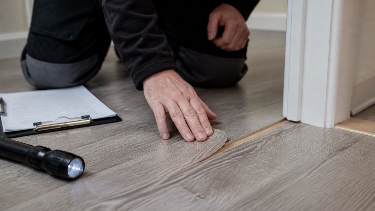Close-up of a technician kneeling on a laminate floor, running a hand over a chipped board edge near a doorway, with a clipboard and torch beside them