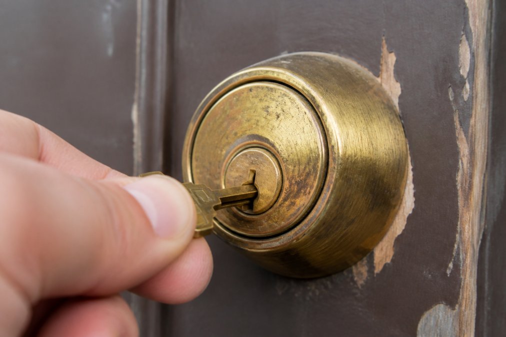 Hand inserting a worn key into a tarnished residential deadbolt that requires lock rekeying due to visible damage.