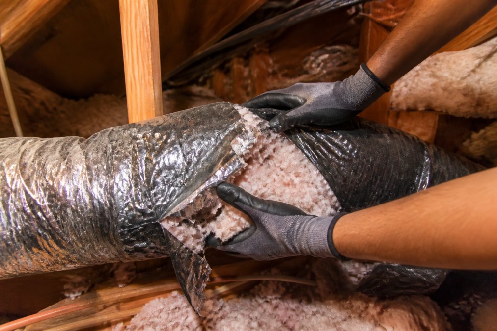 A technician's gloved hands inspecting a torn flexible air duct in a residential attic, dust and loose insulation visible around the damaged section