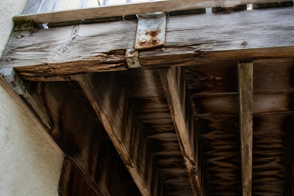 Close-up photograph of a wooden balcony underside showing dark water stains on the joists, cracked and splintered wood beams, and visible corrosion on a metal fastener bracket, lit by harsh afternoon sunlight