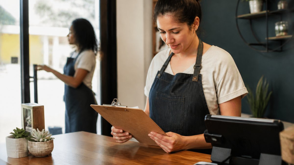 A small business owner at a tidy front desk reviewing a printed opening checklist on a clipboard, a staff member in the background starting a shift, soft morning light through the window