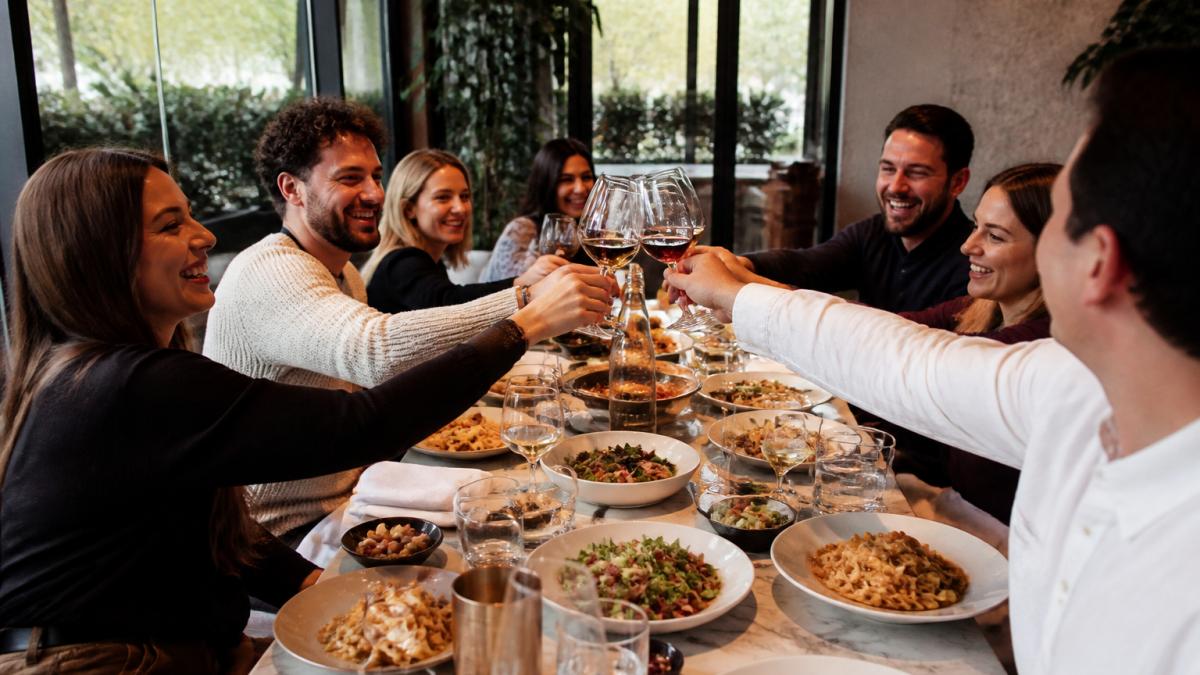 A long table of friends and family sharing handmade pasta and Mediterranean dishes in a warm, stylish restaurant, with glasses raised and natural light from a terrace window
