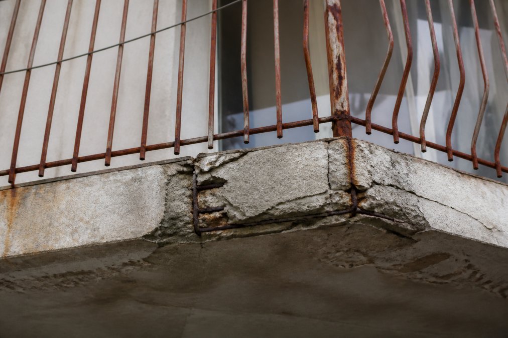 Close-up of deteriorating balcony showing visible structural damage with cracking concrete, rusted fasteners, sagging railings, and water staining indicating poor maintenance and structural compromise