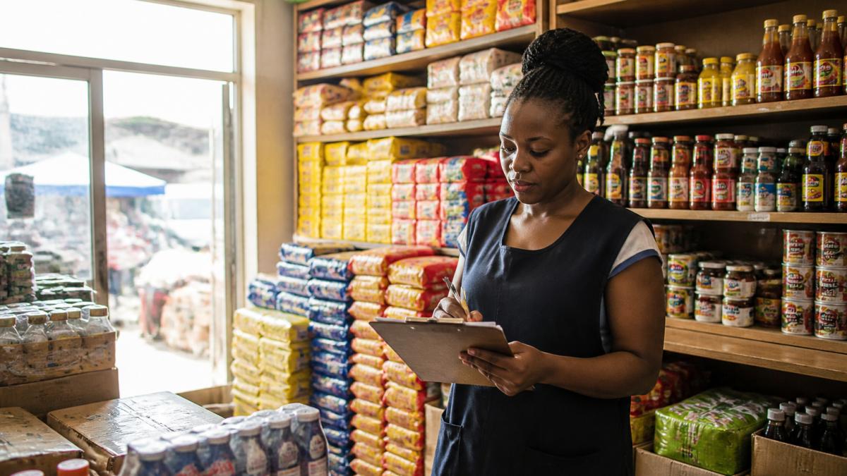 Shop owner using a clipboard to track stock from a wholesale supplier in a sunlit minimart store.