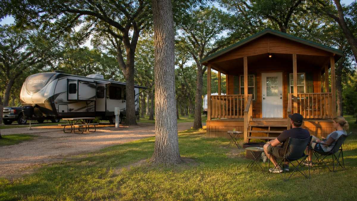 A shaded RV site with full hookups beside a furnished wooden cabin at a wooded Texas resort, late afternoon light filtering through oak trees, a couple relaxing in camp chairs