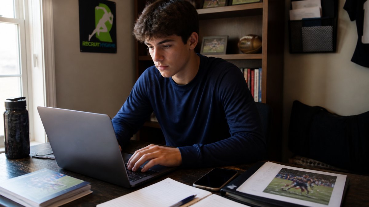 High school student-athlete sitting at a desk with laptop and phone, composing an email to college coaches, focused and determined expression with natural desk lighting
