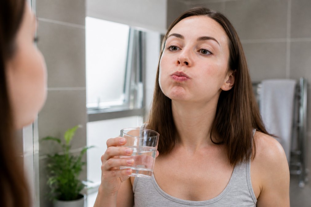 Close-up of a person gently rinsing their mouth with warm salt water in a bright bathroom, holding a small glass, calm and careful expression