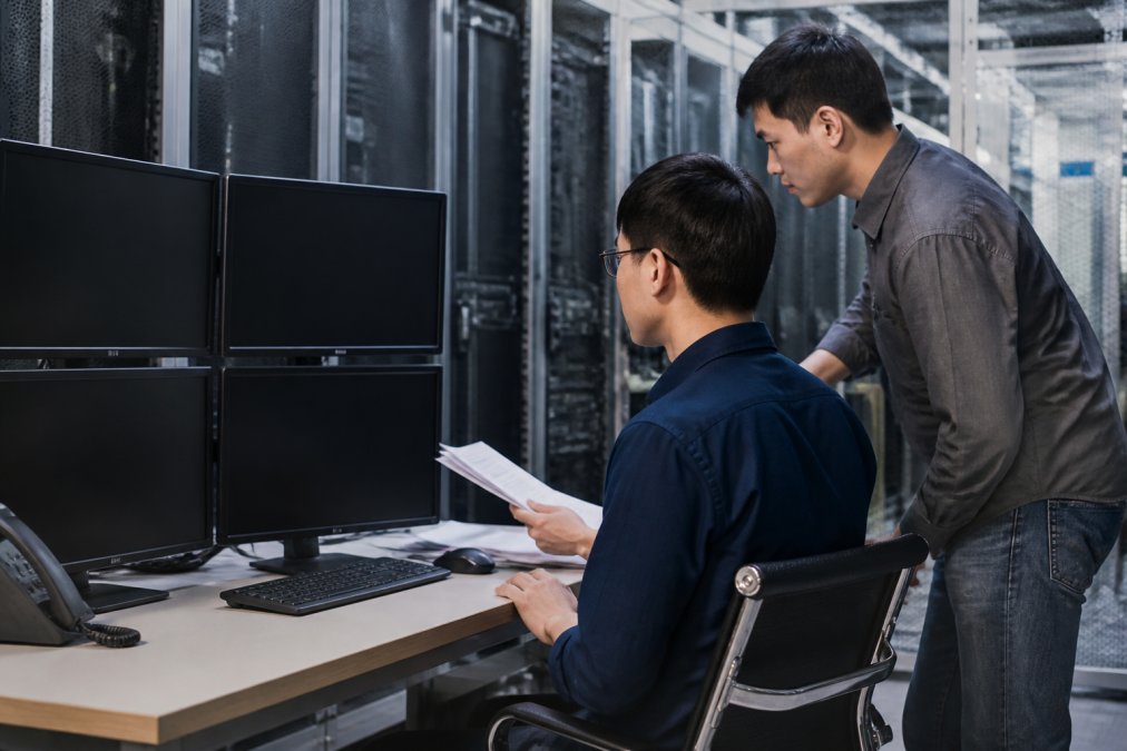IT team members reviewing infrastructure documentation and monitoring dashboards on multiple screens in a modern data center environment with blue-tinted lighting