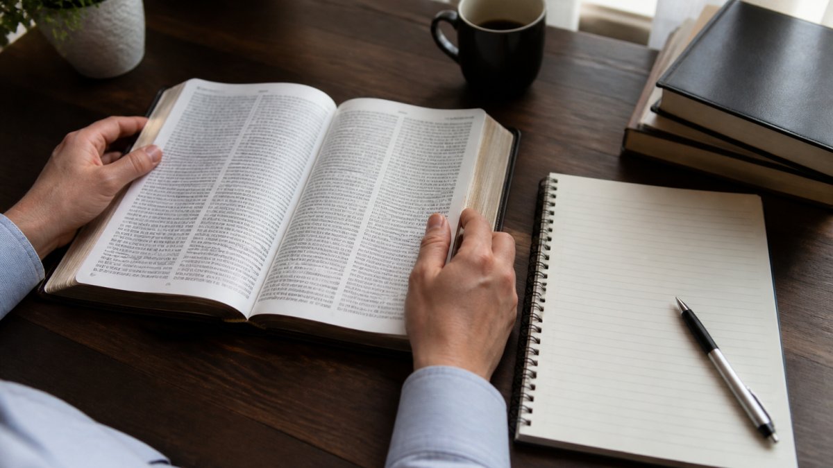 Hands holding an open Bible with a notebook and pen to study Revelation symbolism at a wooden desk