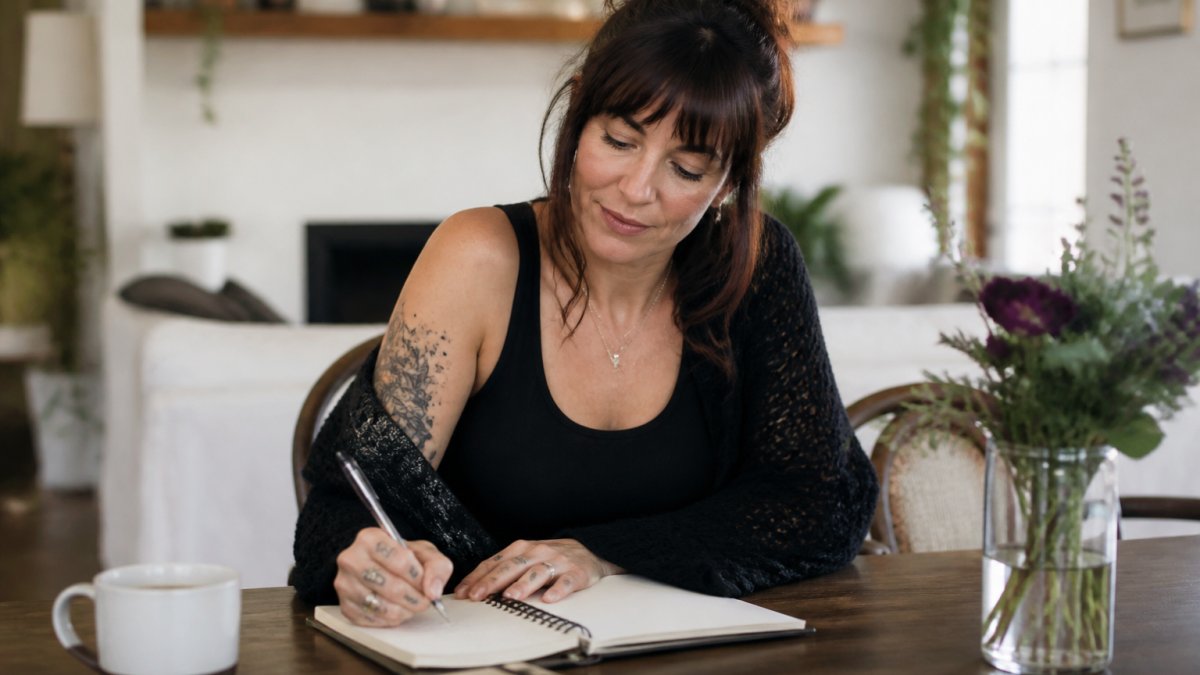 Woman in her 40s sitting at a desk with a notebook, pen, and cup of tea, writing notes about her symptoms in natural daylight