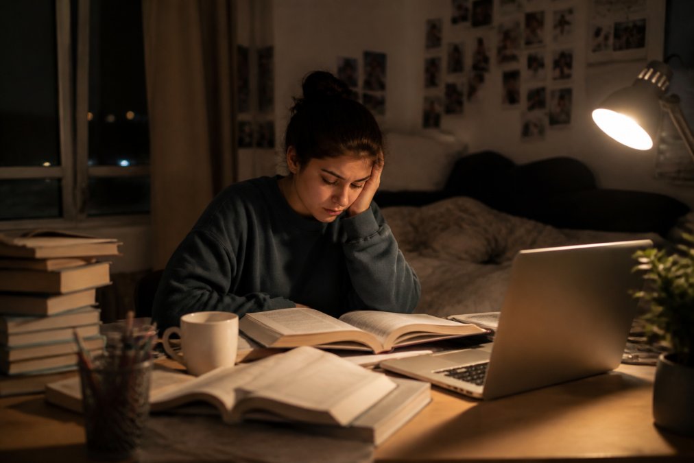 A tired college student sitting at a desk covered in textbooks and a laptop, head resting on one hand, coffee mug nearby, late-night dorm room lighting