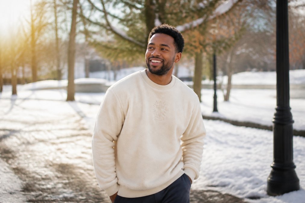 A young adult in a cozy fleece crewneck sweater with a subtle faith-based graphic, smiling while walking through a sunlit winter park with light snow on the ground
