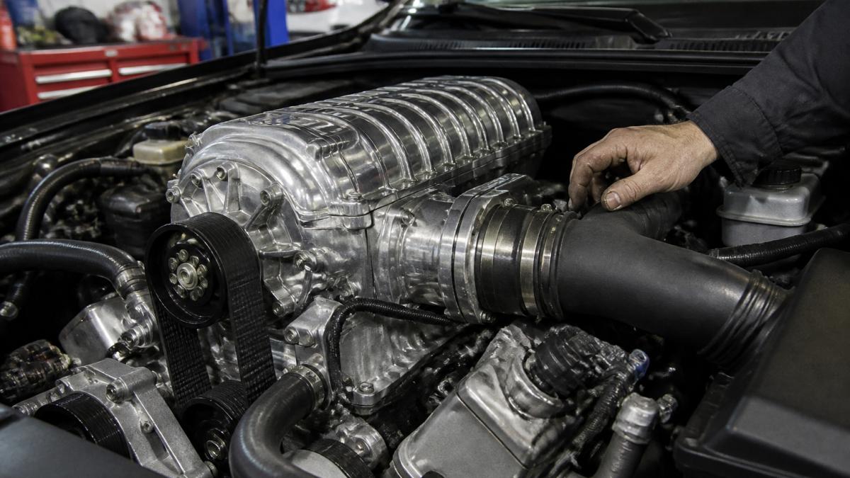 Close-up of a polished twin-screw supercharger unit mounted on an engine, with a mechanic's hand adjusting the intake tube in a well-lit garage