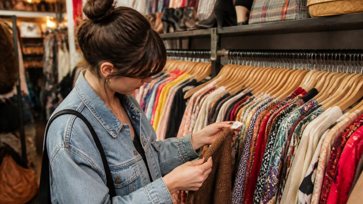 Person browsing racks of vintage clothing and checking fabric quality in a thrift store under warm lighting