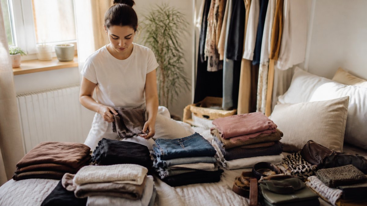 Person sortiert sorgfältig ihre Kleidungsstücke auf einem Bett, mit Stapeln von Oberteilen, Jeans und Accessoires, natürliches Licht von einem Fenster