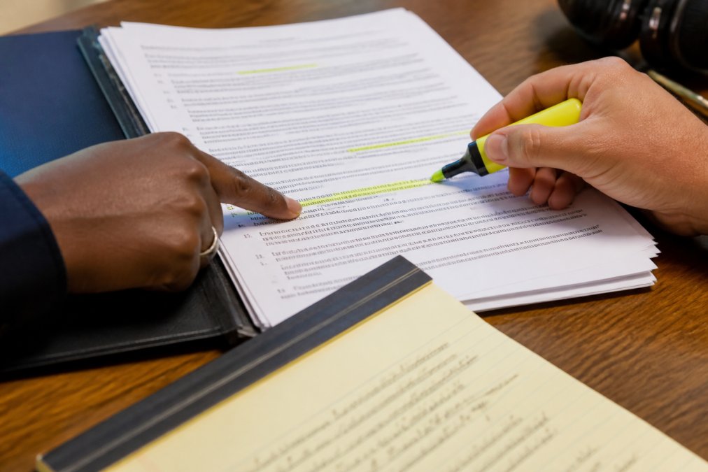 Close-up of hands pointing to specific contract clauses and warning language in a printed music artist agreement, with a highlighter marking key terms and a legal notepad visible on the desk
