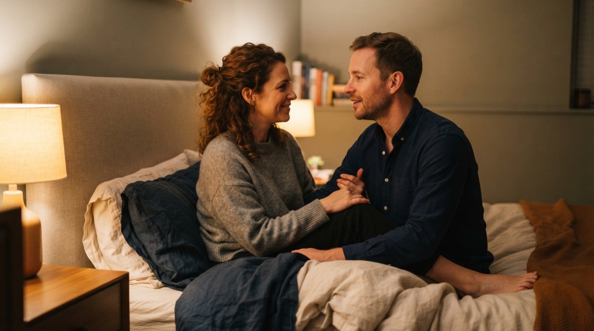Een koppel zit dicht bij elkaar op bed in zachte lampverlichting, ze kijken elkaar aan terwijl ze praten met ontspannen gezichtsuitdrukkingen, wat intimiteit en vertrouwen uitstraalt