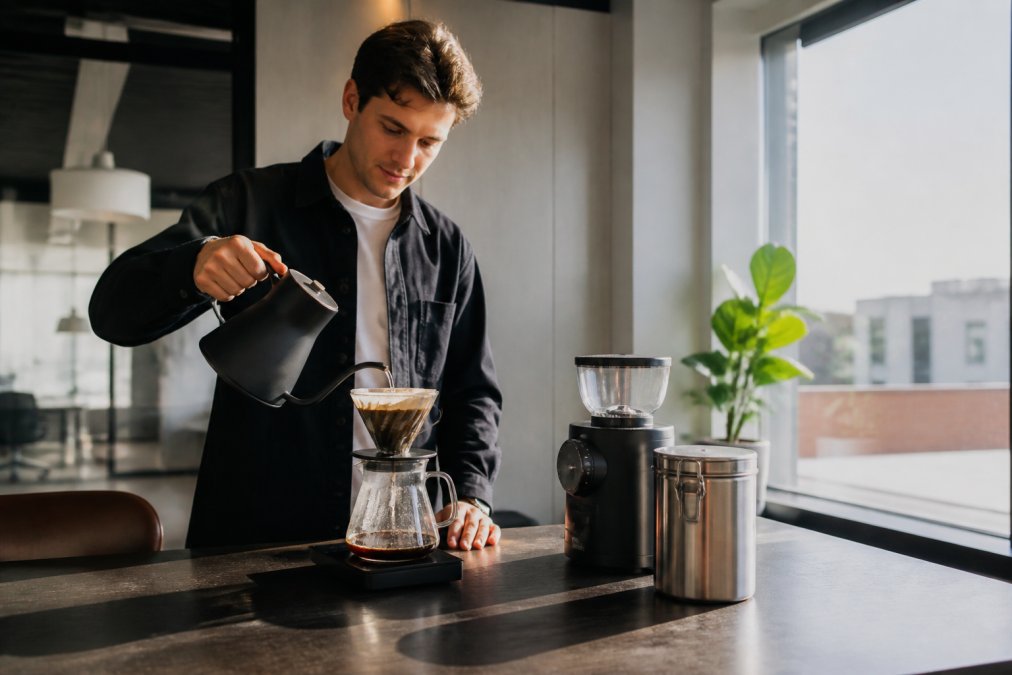 En medarbejder står ved et kontorkøkkenbord og hælder vand fra en elkedel med svanehals over en pour-over dripper, ved siden af en elektrisk kaffekværn og en lufttæt kaffebønnbeholder i rustfrit stål, morgenlys fra et vindue