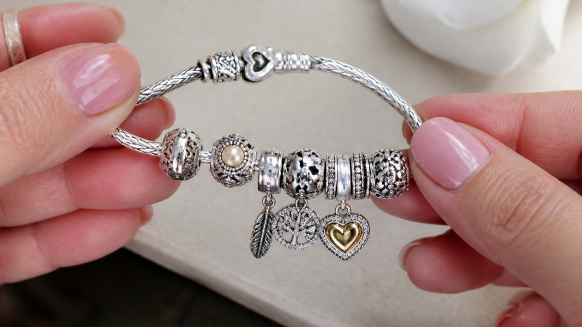 Close-up of a handmade sterling silver charm bracelet being carefully held, showing detailed craftsmanship of individual charm components and intricate clasp work against a soft neutral background with natural lighting
