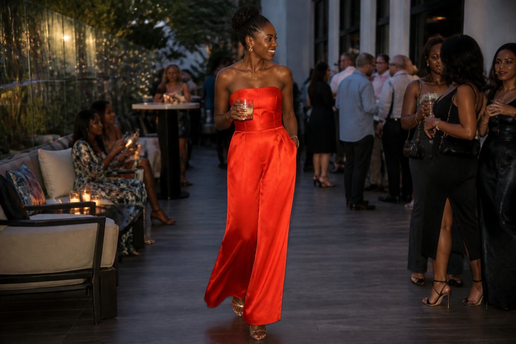 Woman in a flowing red-orange satin wide-leg jumpsuit at an evening formal event, styled with minimal gold accessories and strappy heels, photographed against soft evening lighting