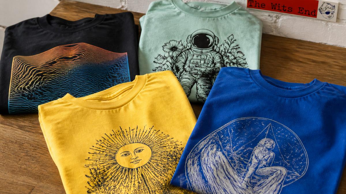 Flat lay of folded progressive graphic tees in various colors (black, mint, yellow, true royal) arranged on a wooden surface, showing design details and fabric quality with natural window lighting