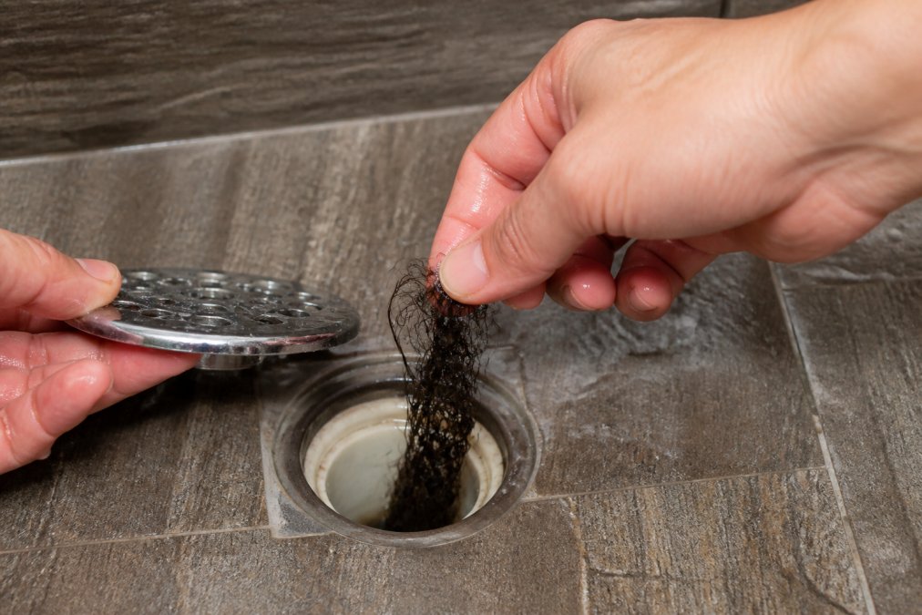 Close-up of hands removing a chrome shower drain cover and extracting a clump of dark hair from the drain opening, with water droplets visible on the metal strainer