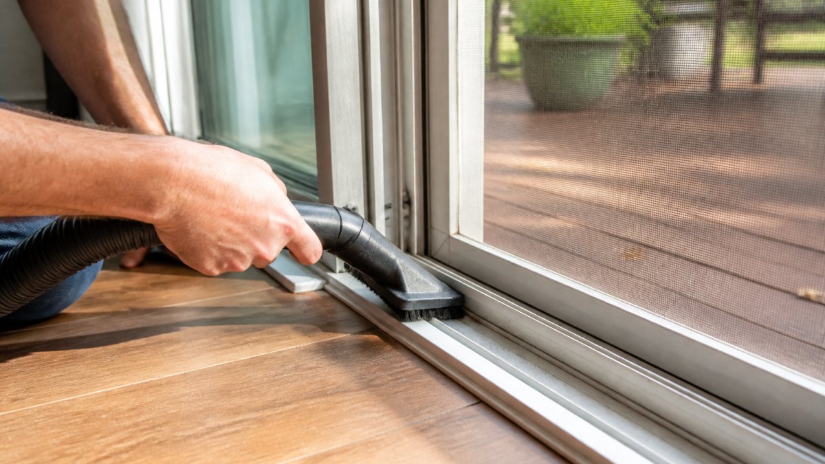 Close-up of a homeowner's hands using a vacuum brush attachment to clear dirt and debris from the bottom track of a sliding screen door, bright daylight through the screen