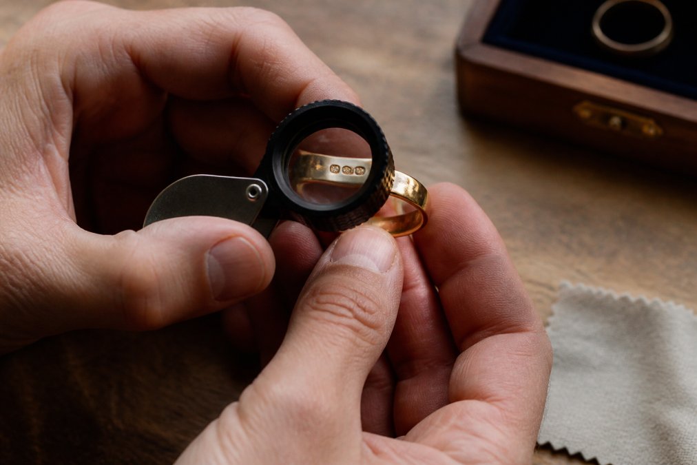 Close-up of hands holding a jeweller's loupe over an antique gold ring, inspecting the hallmark stamps on the band under warm natural light on a wooden table