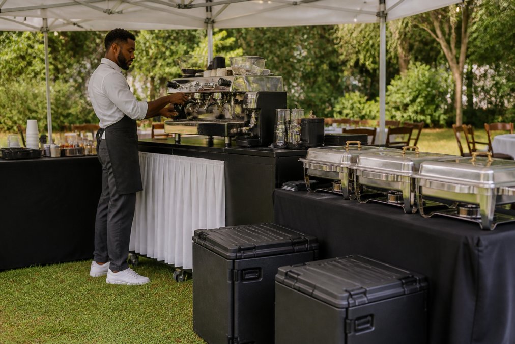Professional barista setting up a mobile coffee cart at an outdoor event with insulated carriers, chafing dishes, and portable serving equipment visible, stainless steel espresso machine gleaming under tent lighting