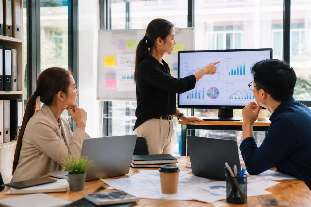 A marketing team in a bright modern office reviewing website visitor analytics on a large monitor, with sticky notes and a whiteboard in the background