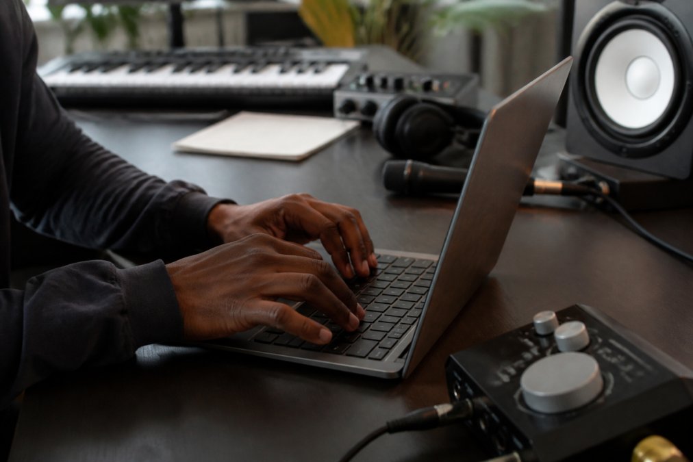 Close-up of a music producer's hands typing on a laptop keyboard in a home studio, with an audio interface and studio monitor visible in the background