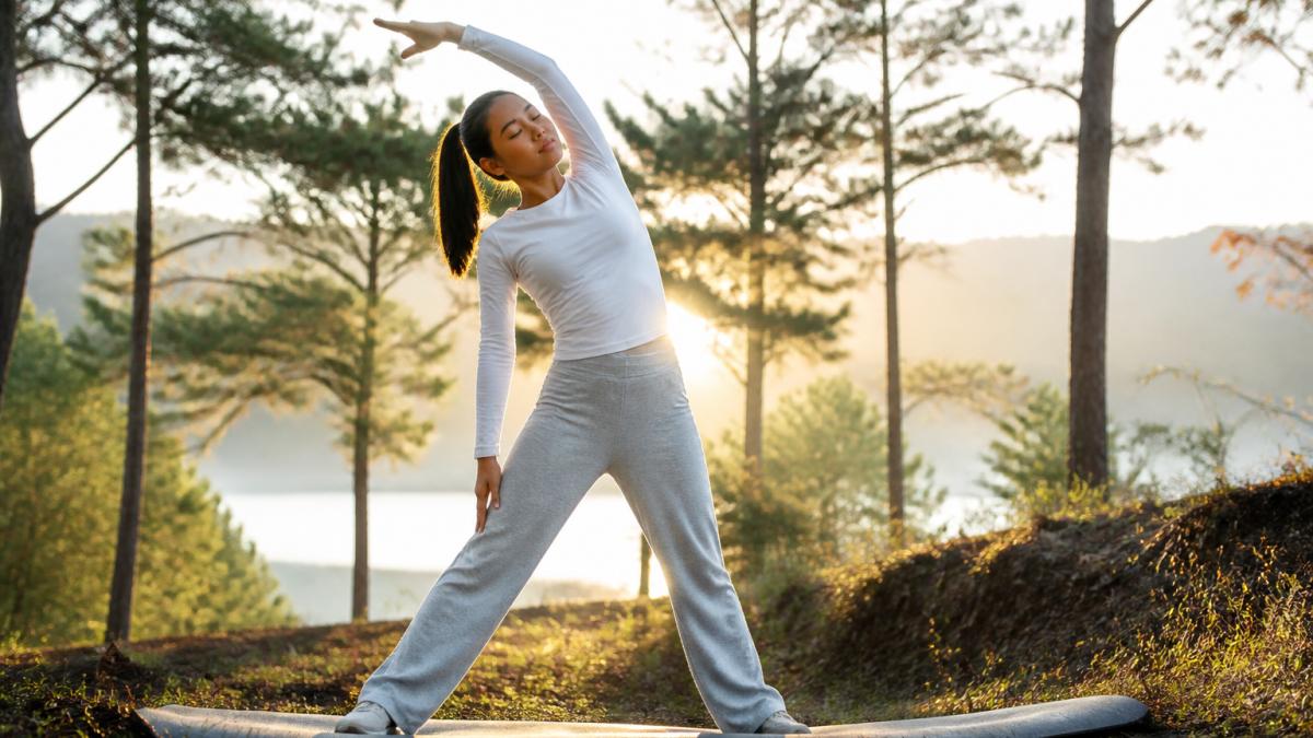 Person performing gentle stretching pose outdoors in natural setting with soft morning light filtering through trees, demonstrating calm, controlled movement